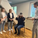 Trener. Pani na krześle testuje google VR. Trzy uczestniczki szkolenia obok.