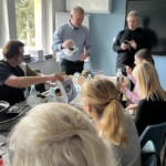 Trener trzyma w ręce gogle VR, zapoznaje uczestników szkolenia z narzędziem. Obok trenera stoi mężczyzna i pobiera na telefon aplikację. Przed nimi przy stołach siedzą uczestnicy szkolenia.