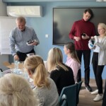 Centralnie stoi trener i kobieta trzymająca gogle VR. Trener instruuje kobietę w obsłudze gogli. Z boku drugi trener z goglami VR w ręce, demonstruje uczestnikom szkolenia kolejne kroki do wykonania.