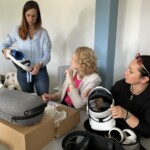 Trenerka stoi i prowadzi instruktaż dopasowywania sprzętu do indywidualnych potrzeb użytkowników. Dwie kobiety zwrócone w stronę trenerki słuchają, jedna z kobiet trzyma w dłoniach gogle. Przed kobietami na stole leżą opakowania i etui na gogle VR.