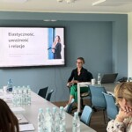 Centralnie tablica multimedialna z prezentowaną treścią zajęć, nieco z boku przed tablicą trenerka warsztatu rozwojowego "Zmiana perspektywy postrzegania" - wprowadzenie do zajęć.